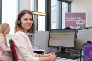 Mitarbeiterin der telefonischen Gesundheitsberatung sitzt am Arbeitsplatz vor einem PC und blickt direkt in die Kamera