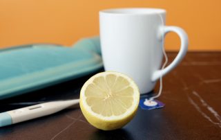 Wärmflasche, Fieberthermometer, Teetasse und eine halbe Zitrone liegen auf einem Tisch