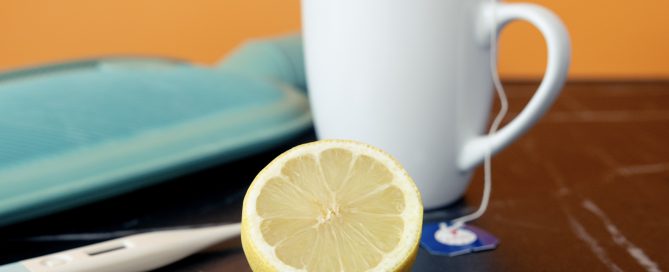 Wärmflasche, Fieberthermometer, Teetasse und eine halbe Zitrone liegen auf einem Tisch